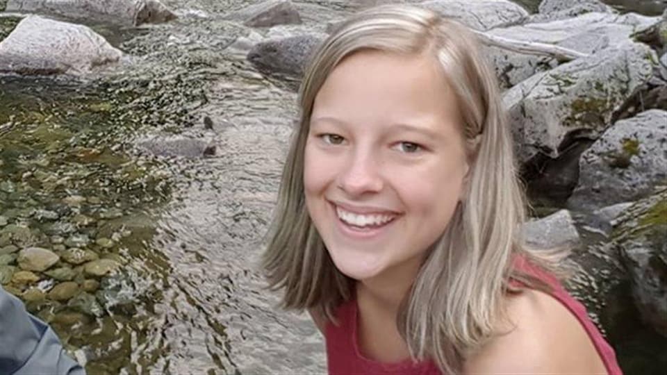 Gros plan du visage d'une jeune fille aux cheveux blonds qui sourit avec, en arrière-plan, un ruisseau et des rochers.