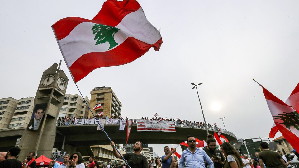 Un homme agite dans les airs le drapeau du Liban. Derrière lui se trouvent des dizaines de personnes, dont certaines sur un pont, qui tiennent des drapeaux libanais et des banderoles. 