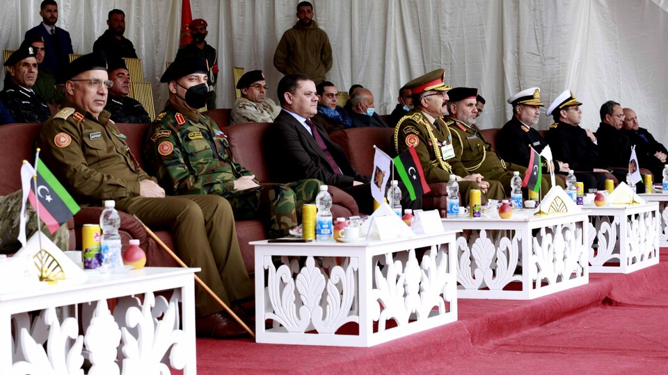 Une série d'hommes, certains en uniforme militaire, sont assis sur des banquettes pour une cérémonie.
