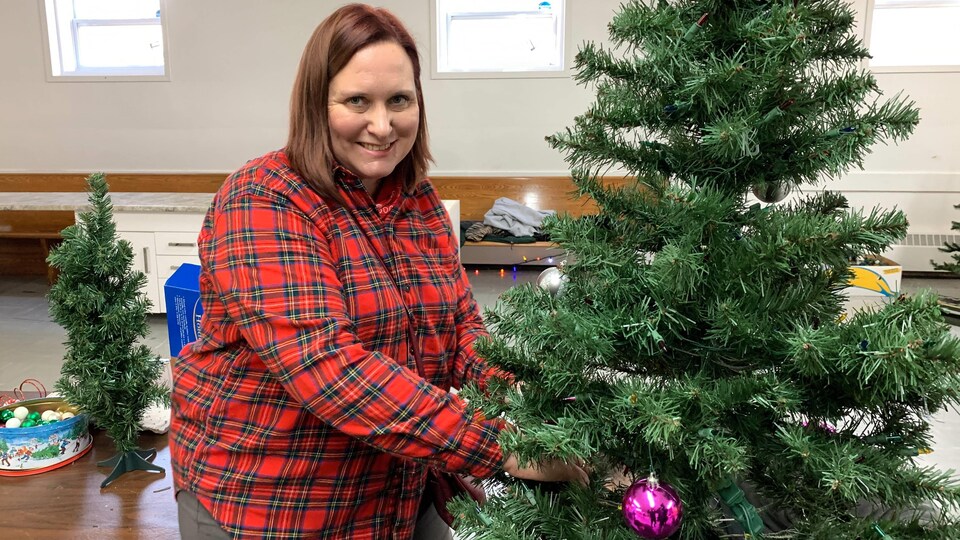 Une femme vêtue d'une chemise à carreaux rouge se tourne vers la caméra et sourit pour une photo. Elle est en train de décorer un sapin de Noël.