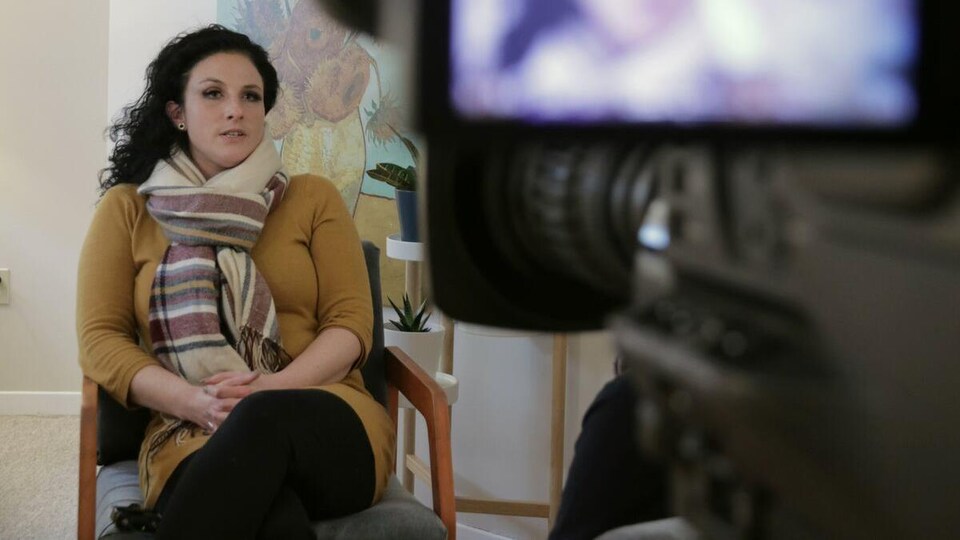 Une femme assise sur une chaise est filmée par une caméra pendant qu'elle accorde une entrevue.