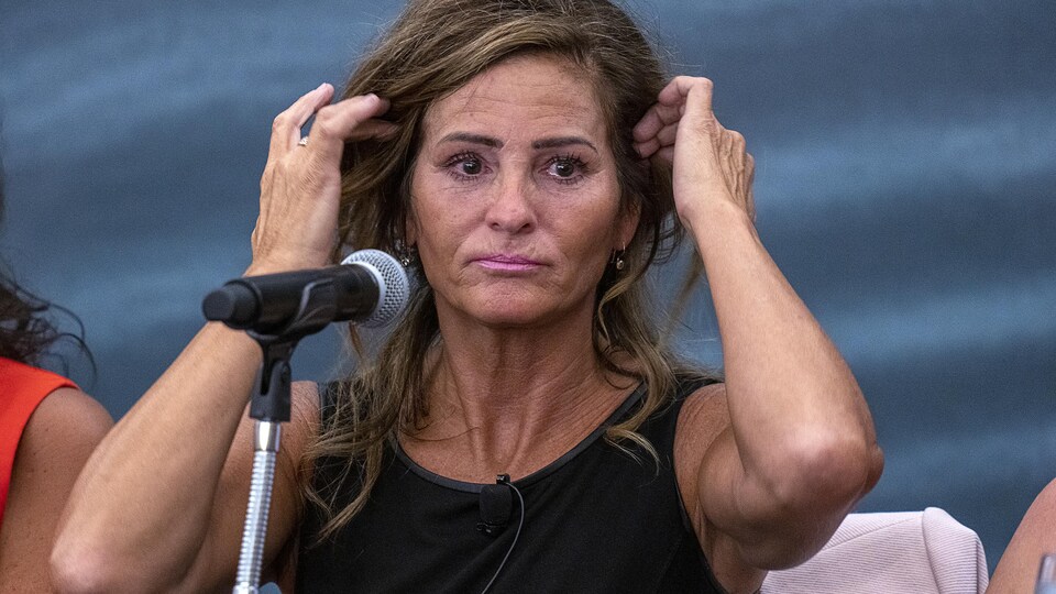 Assise devant un micro, Lisa Banfield lève ses deux mains pour replacer ses cheveux.