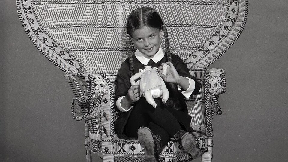 Une fillette portant des tresses, assise sur une grande chaise en paille, tient un toutou dans ses mains.