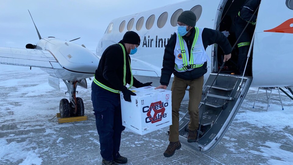 Deux hommes portant des masques chirurgicaux transportent une boîte à la sortie d'un petit avion.