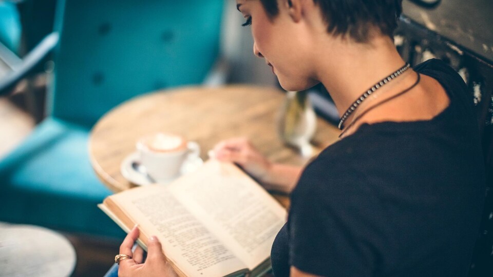 Une jeune femme vue de dos lit un livre dans un café.