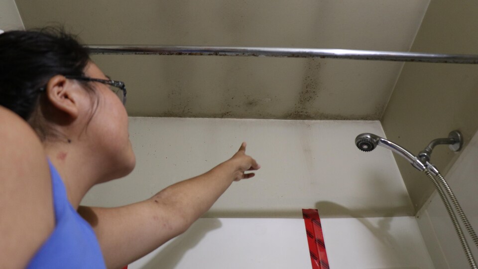 Une femme dans une salle de bain montre des moisissures sur le plafond et le mur.