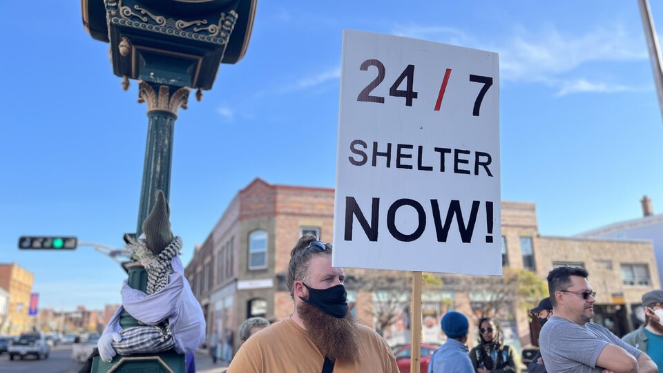 Un groupe dehors devant l'hôtel de ville. Un homme brandit une affiche sur laquelle on lit « Des abris 24 heures et 7 jours maintenant ».