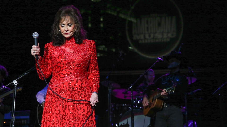 Loretta Lynn porte une robe rouge et tient un micro sur une scène.