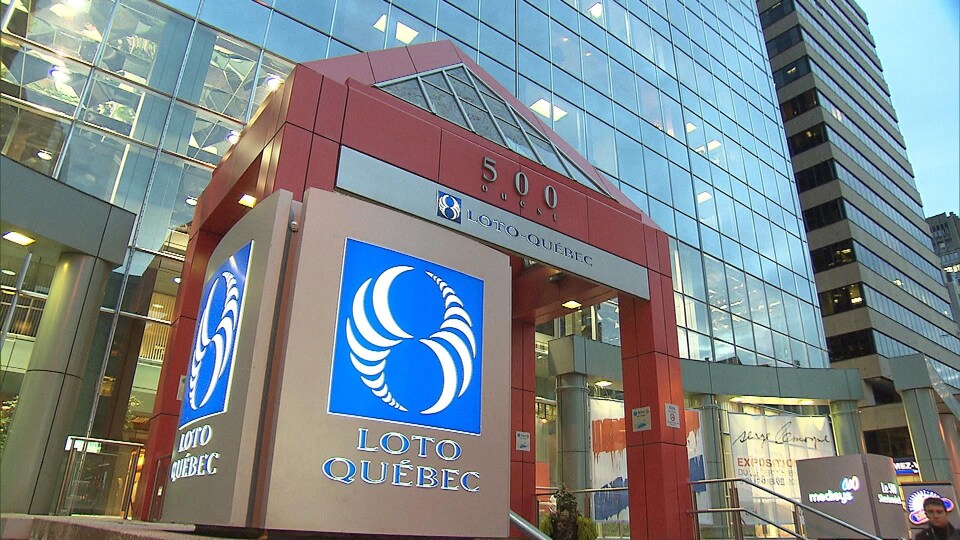 Un logo circulaire bleu et blanc, sous lequel on peut lire Loto Québec, est photographié devant un gratte-ciel.