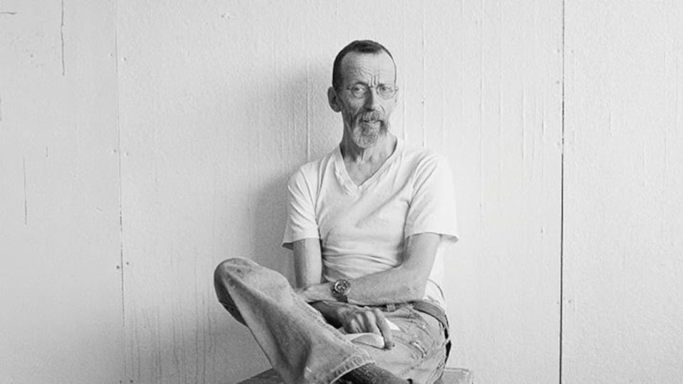 L'homme vêtu d'un t-shirt blanc est assis sur un tabouret, les jambes croisées, devant un mur blanc.