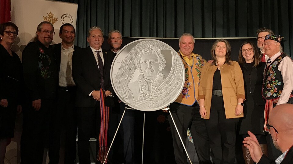 Des Métis, des hommes politiques et des représentants de la Monnaie royale canadienne posent derrière la nouvelle pièce commémorative d’un dollar à l'effigie de Louis Riel.