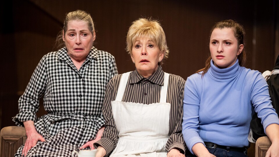 Trois personnages féminin sont assis sur un vieux divan avec un air effrayé. 