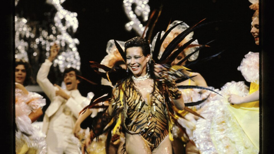 Louise Forestier, souriante, porte un costume extravagant sur scène avec danseurs en arrière-plan.