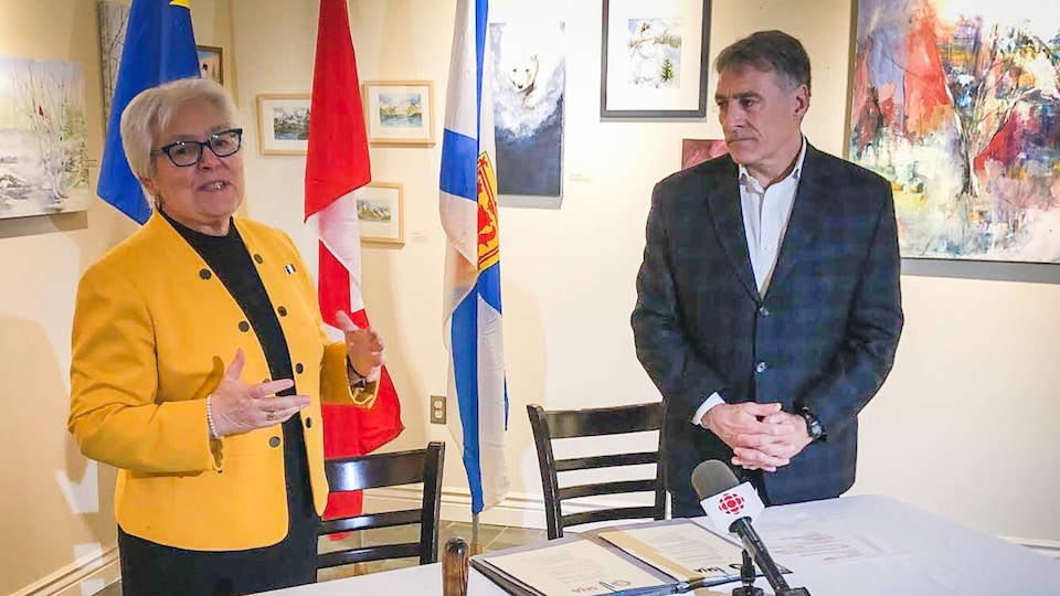 Louise Imbeault et Allister Surette derrière une table avec une nappe sur laquelle sont posés des micros de journalistes et devant des drapeaux.