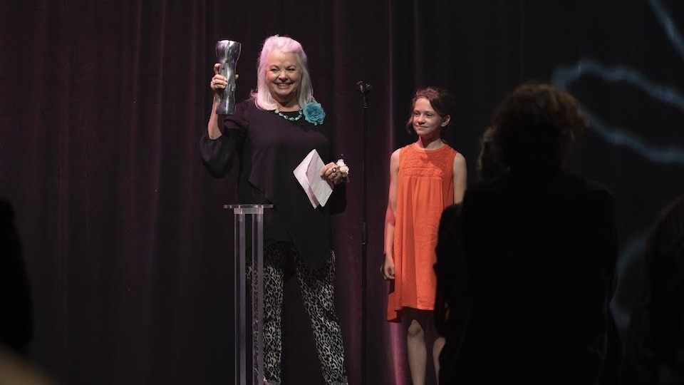 A woman is holding a trophy on a stage, along with a child wearing an orange dress. 