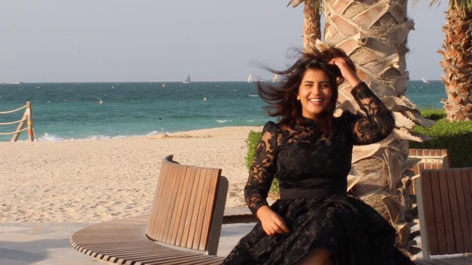 Une femme assise sur un banc à la plage près de l'eau. Elle a les cheveux dans le vent et porte une robe noire. 