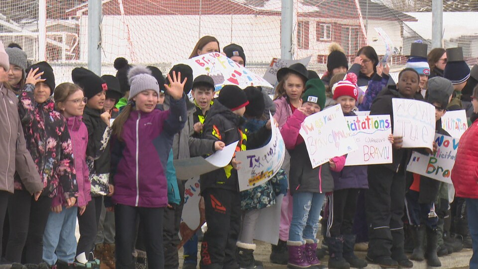 Des enfants tiennent des pancartes félicitant Luc LeBlanc et le saluent alors qu'il passe devant eux en autobus. 