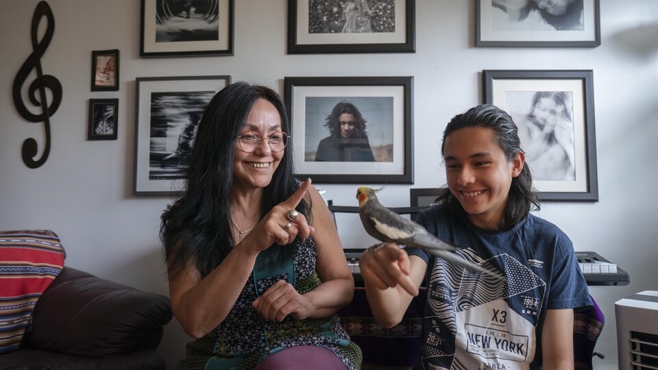 Une mère et son fils adolescent, qui a un oiseau perché sur le doigt, sourient en regardant l'oiseau, dans leur appartement.