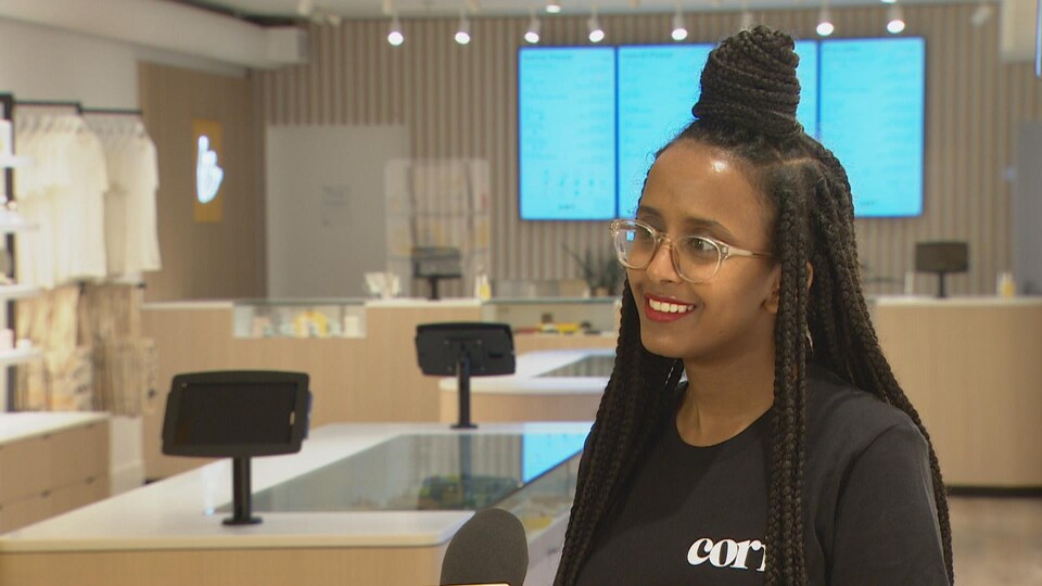 Une jeune femme à l'intérieur d'une boutique de cannabis. Derrière elle, des comptoirs et des panneaux numériques.