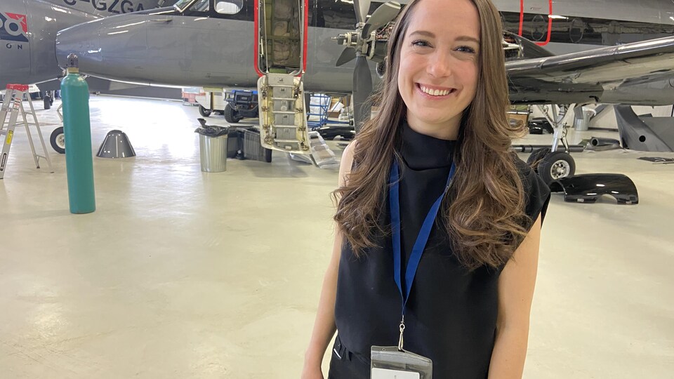 Une femme sourit devant un avion dans un hangar.