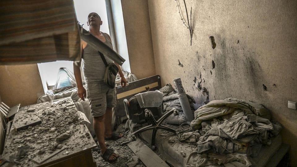 A man examines the damage in a room of his house. A rocket is embedded in the mattress of a bed.