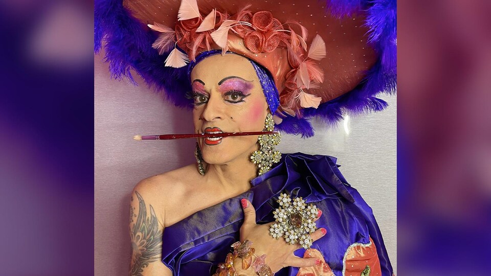 Une drag queen tient un pinceau dans sa bouche maquillée.