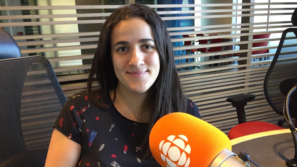 Une femme souriante aux cheveux longs noirs est assise dans un studio de Radio-Canada-Windsor. Devant elle il y a un micro de Radio-Canada