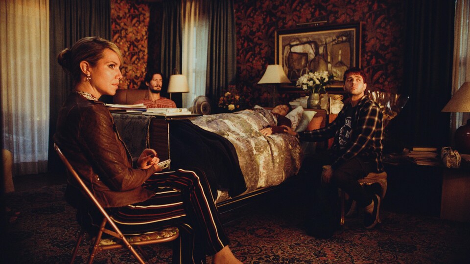 Une femme et deux hommes sont assis autour d'un lit où est couchée une femme plus vieille. 