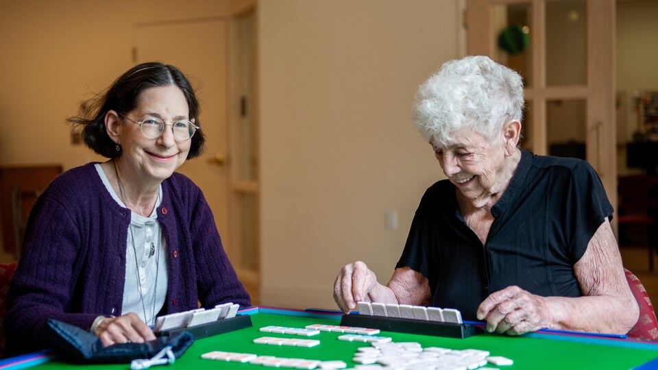 Deux aînés jouent au mah-jong en souriant. Plusieurs tuiles du jeu sont posées sur la table tandis que l'une d'elle prépare ses combinaisons et que l'autre sourit à la caméra.