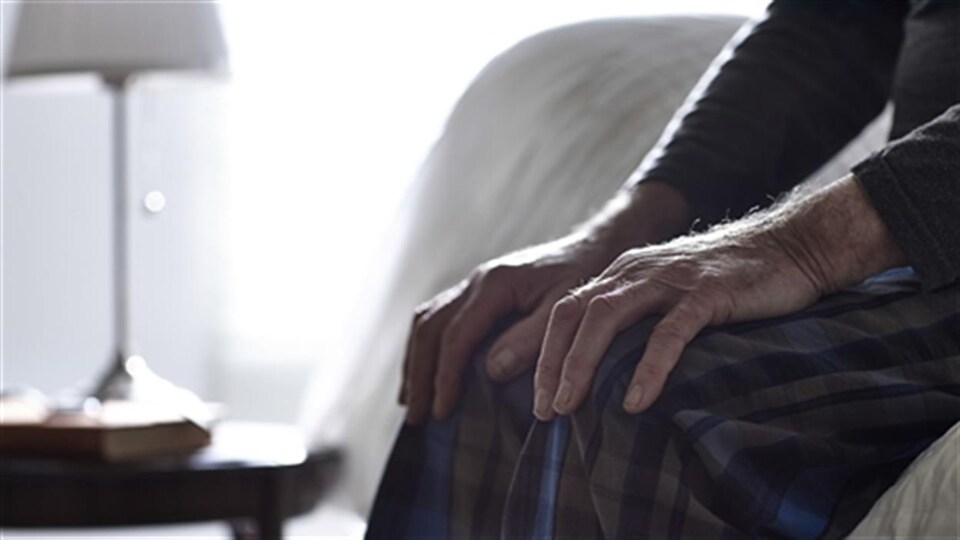 Un homme âgé assis sur son lit, avec les mains sur ses genoux, alors qu'on voit une lampe en arrière-plan.