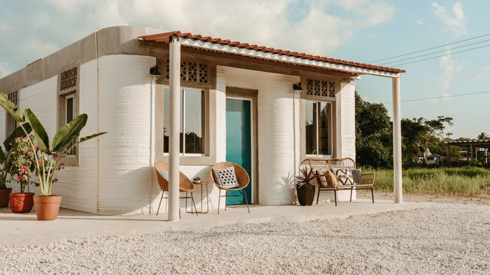 Une maison de béton imprimée en 3D entourée de plantes et de meubles de jardin. 