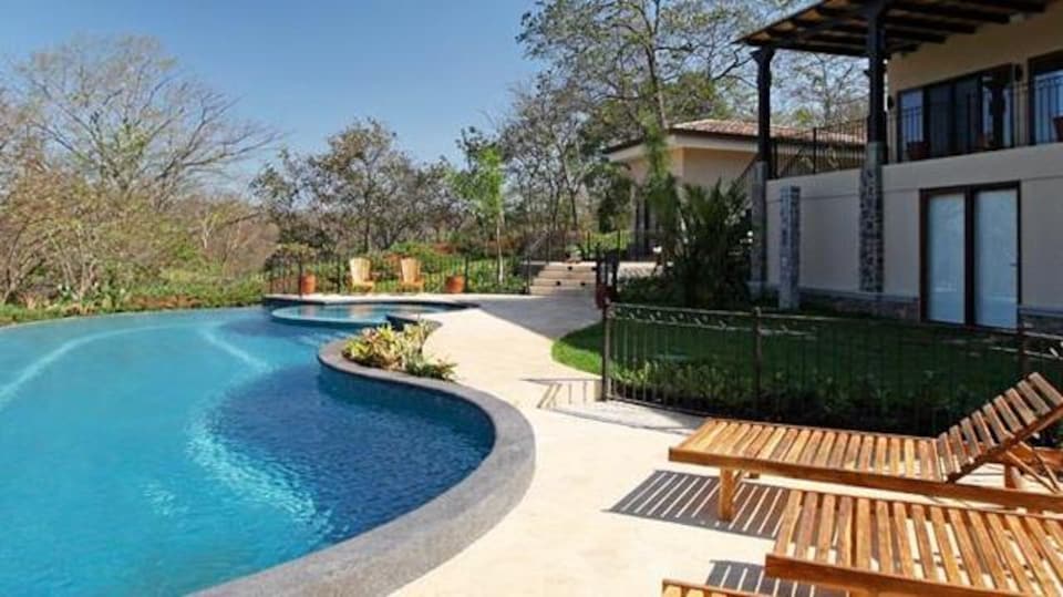 Vue d'une piscine bordée de transats en bois et d'une maison aux murs clairs.
