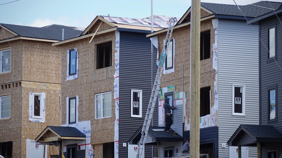 Un travailleur debout sur le portique d'une maison en construction est en train d'installer du revêtement extérieur.