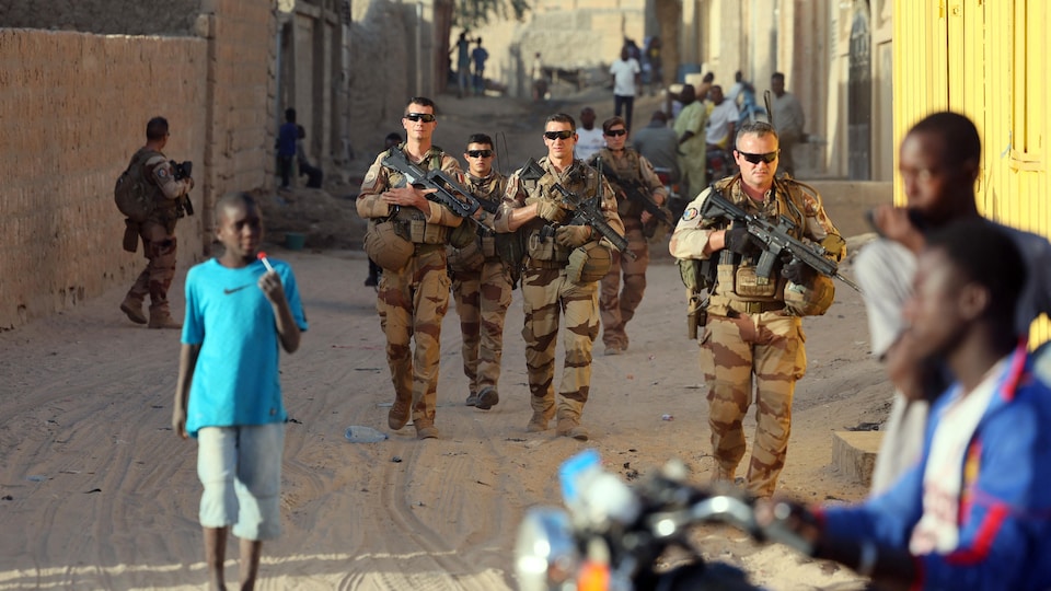 Des soldats en uniforme dans une rue, où on voit aussi des passants.