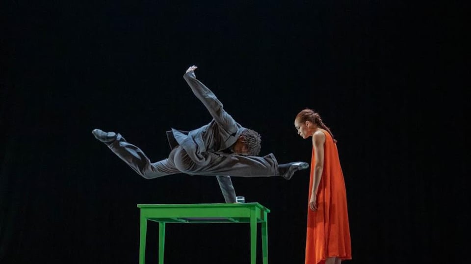 A man dressed in a suit balances his hand on a green table, while a woman in an orange dress looks on. 