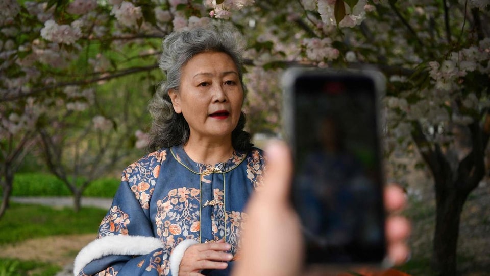 Une femme dans un parc avec des arbres en fleur parle devant un téléphone cellulaire qui la filme.