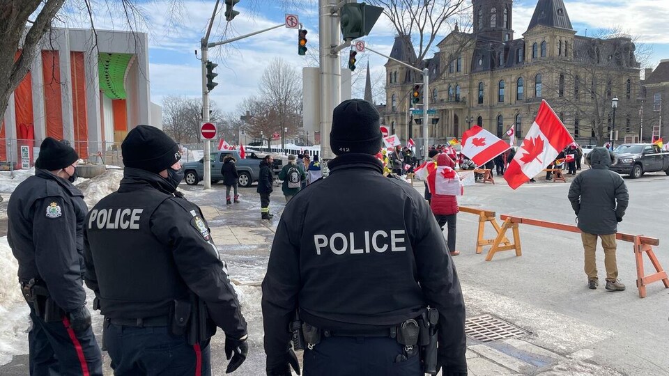 Trois policiers de dos observent un petit groupe de manifestants avec des drapeaux du Canada près de l’Assemblée législative du Nouveau-Brunswick.