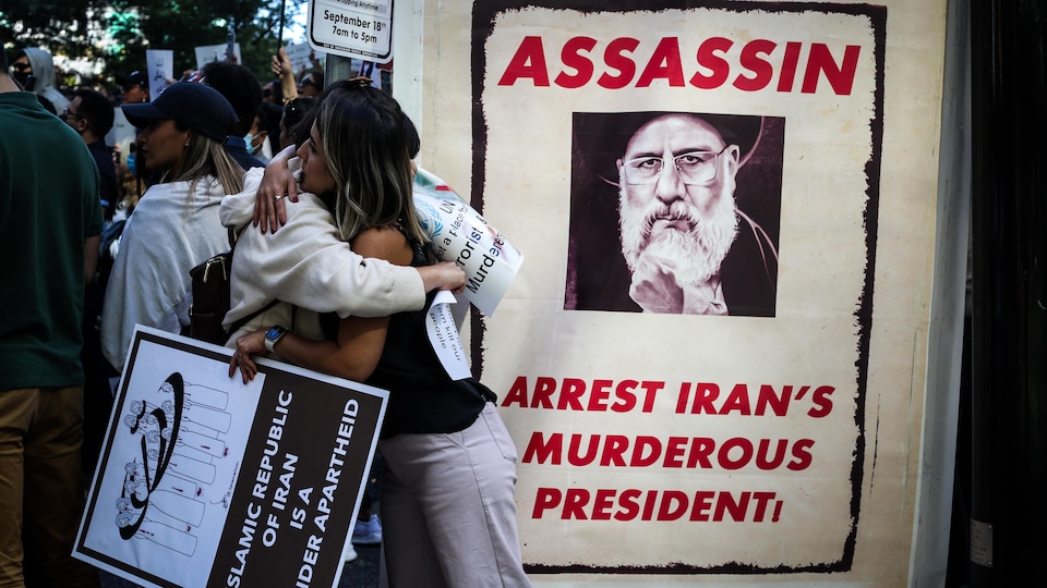 Deux manifestants se prennent dans les bras, derrière une pancarte du président iranien où il est écrit assassin. 