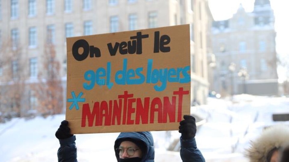 Un homme tient une pancarte marquée du slogan « On veut le gel des loyers maintenant ».