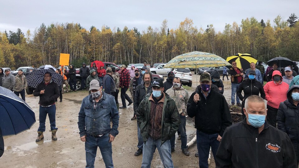 Des hommes et quelques femmes au visage masqué, certains tenant un parapluie, se tiennent debout dans une clairière.