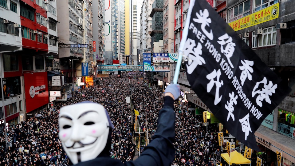 Un manifestant portant un masque de Guy Fawkes brandit un drapeau devant une foule en contre-bas.