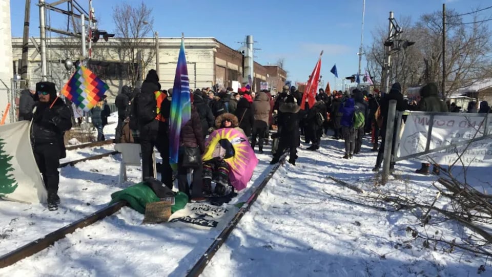Des manifestants sont rassemblés sur des voies ferrées enneigées avec des drapeaux et des pancartes.