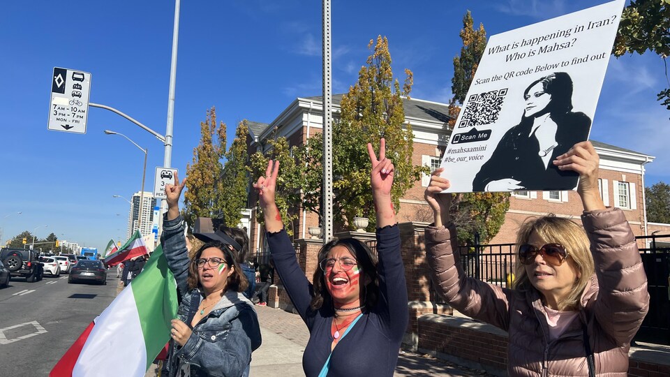 Trois femmes sur le trottoir montrent des signes de paix avec leurs mains. L'une tient une affiche de Mahsa Amini et une autre tient un drapeau iranien.