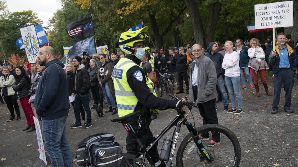 Un policier à vélo et qui porte un masque, au milieu de manifestants qui protestent contre le port obligatoire du masque.