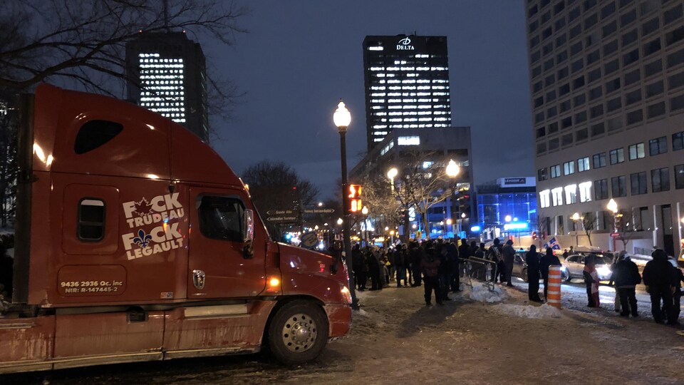Des manifestants en bordure du boulevard René-Lévesque. Au premier plan, un tracteur semi-remorque rouge sur lequel on peut lire le mot en f devant les noms des premiers ministres Trudeau et Legault.