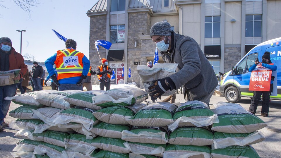 Un homme dépose un sac de sable sur d'autres sacs érigés en mur, alors que des personnes aux couleurs de l'APTS manifestent.