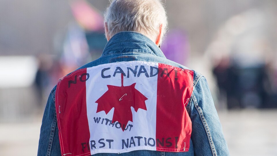 Sur le manteau de l'homme se trouve un drapeau du Canada à l'envers sur lequel on peut lire « Pas de Canada sans Premières Nations ».