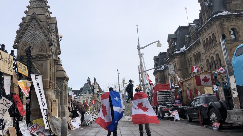 Des manifestants sur un trottoir, de dos, avec des drapeaux du Canada et du Québec.