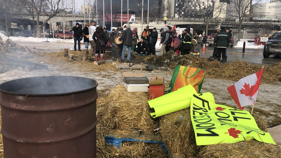 Des manifestants sont réunis autour d'un feu.. Ils ont des tambours autochtones dans les mains. Au premier plan, des pancartes de manifestants. 