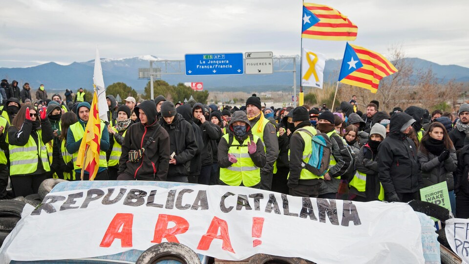 Plusieurs centaines de personnes bloquent une autoroute. Certains sont vêtus de noir ou de dossards jaunes. On voit le drapeau catalan qui flotte dans la foule. 